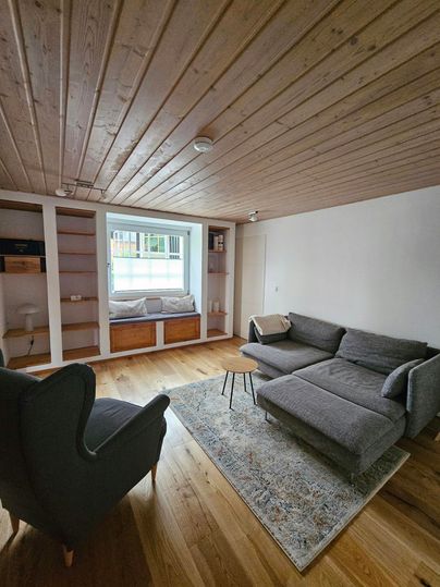 Een moderne woonkamer met een grijze bank en een comfortabele fauteuil. Grote ramen laten veel licht binnen en er zijn lichte houten meubels.