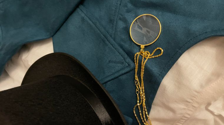 Ein eleganter schwarzer Zylinderhut liegt neben einer blauen Weste. Dazu gehört eine goldene Kette mit einer runden Lupe.