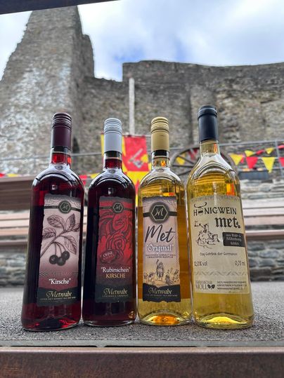 Vier flessen verschillende soorten mede staan op een tafel. Op de achtergrond is een middeleeuwse kasteelruïne en kleurrijke vlaggen te zien.
