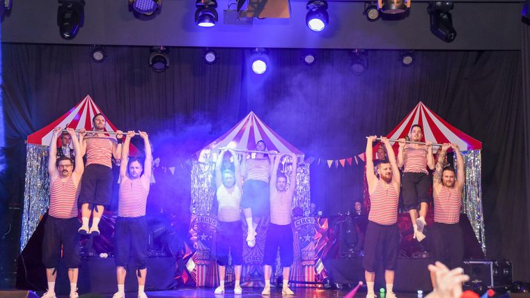 Een groep artiesten voert een acrobatische act op een podium op. Op de achtergrond zijn circus tenten en gekleurde lichten te zien.