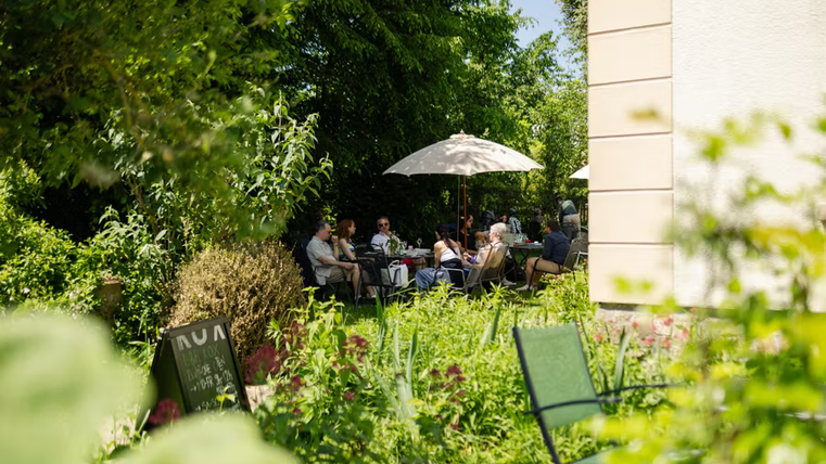 A sunny garden with guests sitting under sun umbrellas. Surrounded by many plants and an inviting atmosphere.
