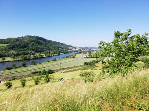 Een pittoresk landschap met een rivier, omgeven door glooiende heuvels en groene weiden. De lucht is helder en blauw, wat de idylle versterkt.