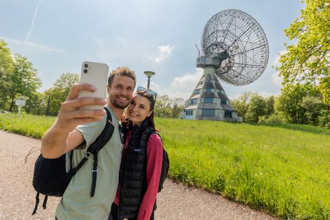 Un couple prend un selfie devant le radiotélescope Astropeiler Stockert dans un paysage verdoyant.