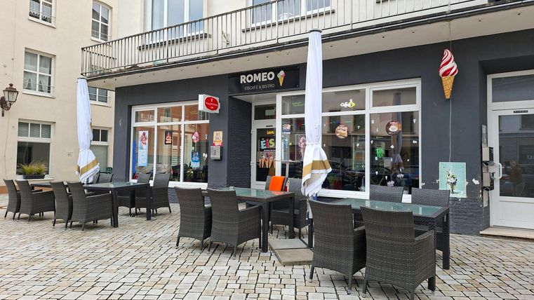 Ein Café mit Außensitzplätzen und Sonnenschirmen. Das Gebäude hat große Fenster und eine einladende Atmosphäre.