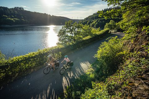 Deux cyclistes roulent sur un chemin le long d'un lac sous le soleil, entourés d'une nature verdoyante.