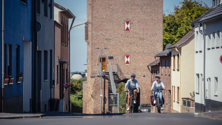 Deux cyclistes roulent sur une route à Zülpich, avec la porte de Bachtor en arrière-plan.