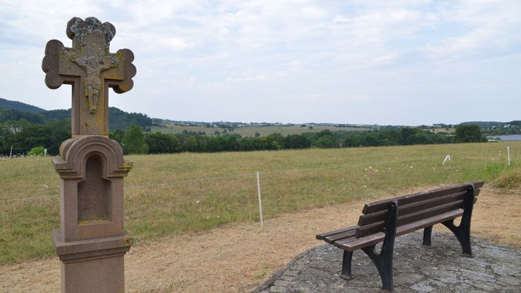 Un banc et une croix en pierre dans un paysage rural avec une vue étendue sur les champs et les collines.