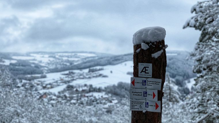 Besneeuwd wandelpad met wegwijzer op de voorgrond en winters landschap op de achtergrond.