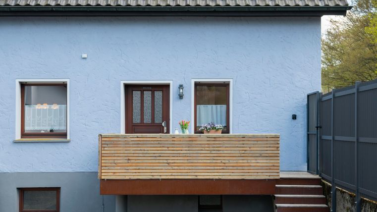 Een blauw huis met een houten balkon en een voordeur. Naast de ramen staan bloempotten met planten.