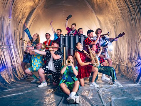 Un groupe de musiciens pose dans des costumes colorés dans une pièce scintillante. Ils tiennent divers instruments de musique et dégagent de la joie.