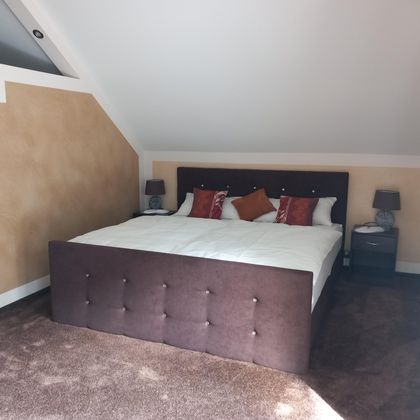 A cozy bedroom with a large double bed and decorative cushions. The walls are painted in warm colors, and there are nightstands with lamps.