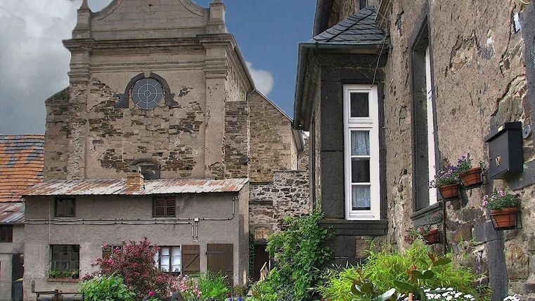 Ein altes, charmantes Gebäude mit einer markanten Fassade steht neben einem gepflegten Garten. Bunte Blumen und Pflanzen verleihen der Umgebung einen einladenden Charakter.
