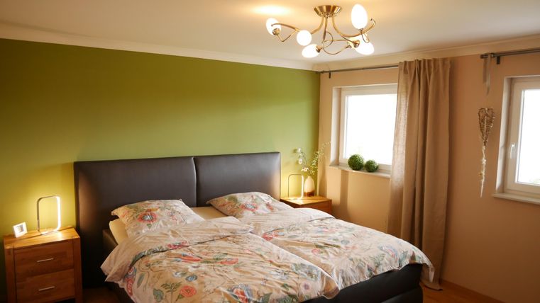 Een gezellige slaapkamer met een tweepersoonsbed en kleurrijke dekbedovertrekken. Aan de muren zijn groene en beige tinten, en de ramen laten veel daglicht binnen.