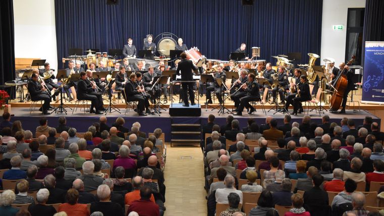 An orchestra is playing on a stage in front of a full audience. The conductor stands in the center and directs the musicians.