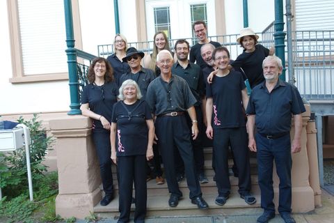 Eine Gruppe von Personen steht auf einer Treppe vor einem Gebäude. Sie tragen schwarze Kleidung und lächeln in die Kamera.