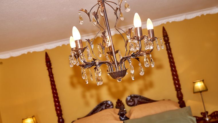 Ein elegantes Schlafzimmer mit einem Kronleuchter und einem großen Bett. Die Wände sind in warmem Gelb gestrichen und es gibt sanfte Beleuchtung von Nachttischlampen.