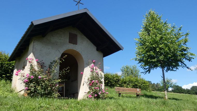 Petite chapelle dans la verdure avec des roses en fleurs et un banc à côté.