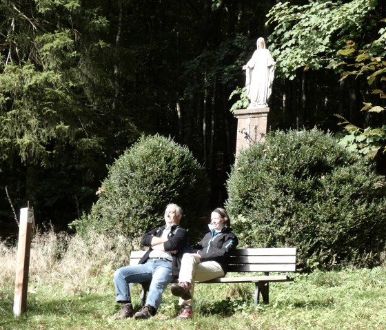 Two people are sitting on a bench in front of a Marian column in the forest. They are enjoying the sun, surrounded by bushes and trees., © TI Bitburger Land