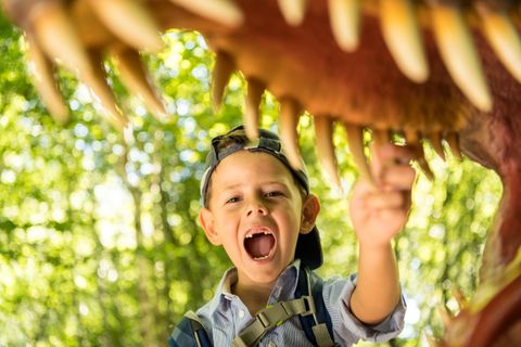 A boy stands under a dinosaur model with his mouth open and points to his teeth. Trees can be seen in the background.