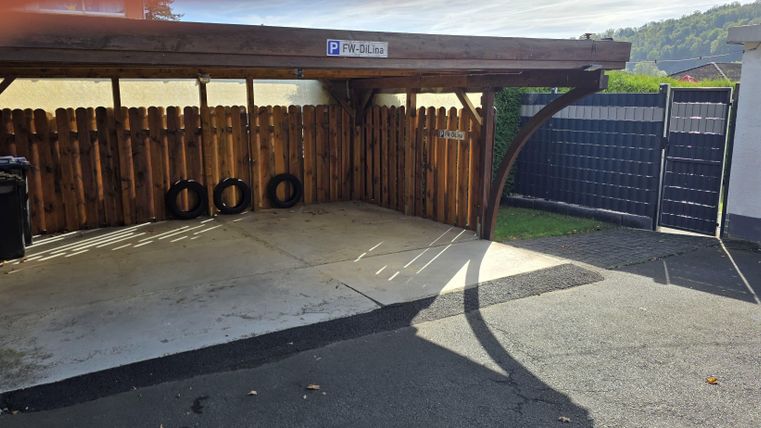 A covered parking area with wooden walls and an entrance gate. In the background, tires and trash bins are visible.