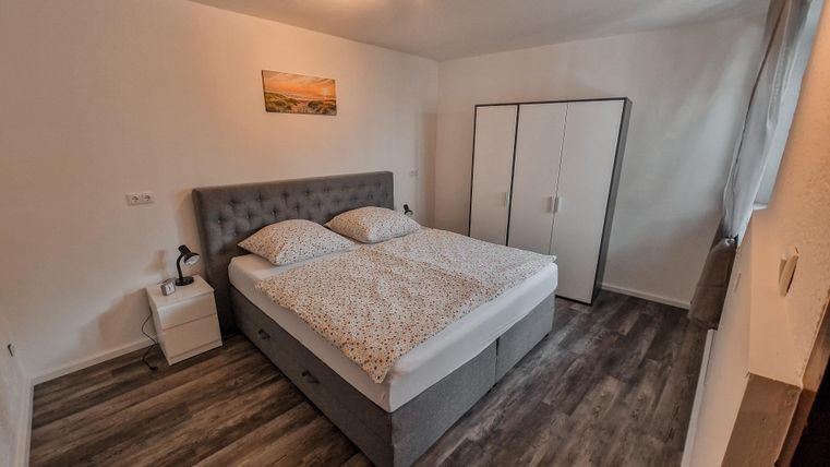 Ein modernes Schlafzimmer mit einem großen Doppelbett und hellen Wänden. Es gibt einen Schrank und ein Fenster, das Tageslicht hereinlässt.