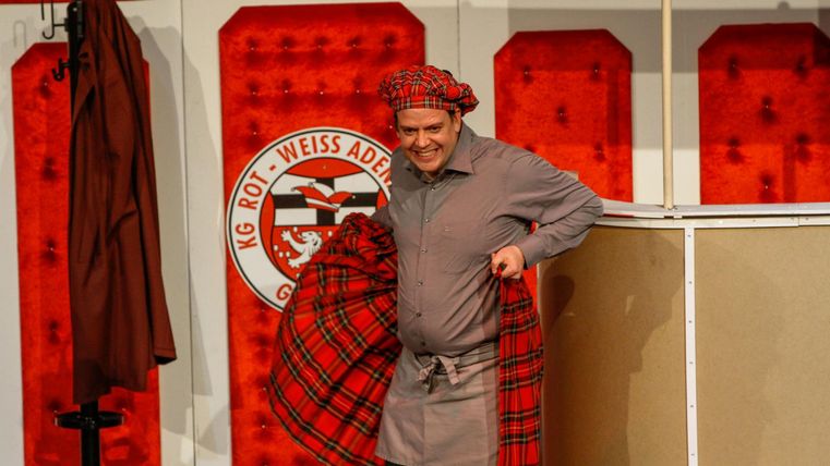 A man in traditional clothing with a checked pattern stands on the stage. In the background, red walls and clothing racks can be seen.