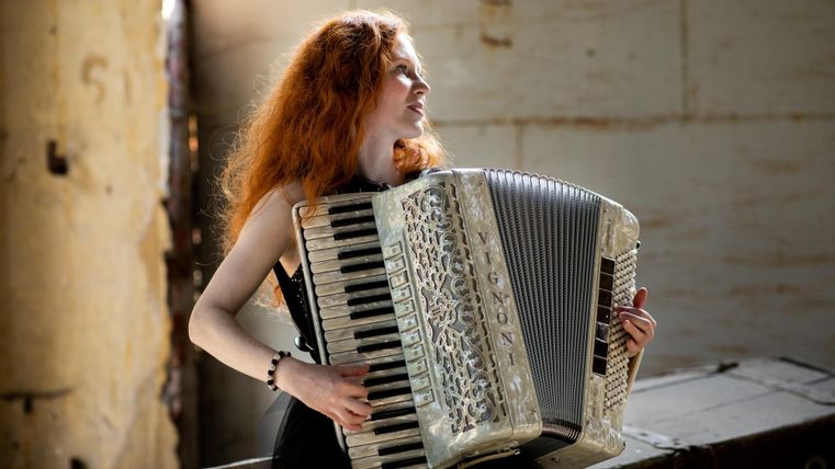 Ein Mädchen mit roten Locken spielt Akkordeon in einem rustikalen Raum. Das Licht fällt sanft auf sie und schafft eine gemütliche Atmosphäre.