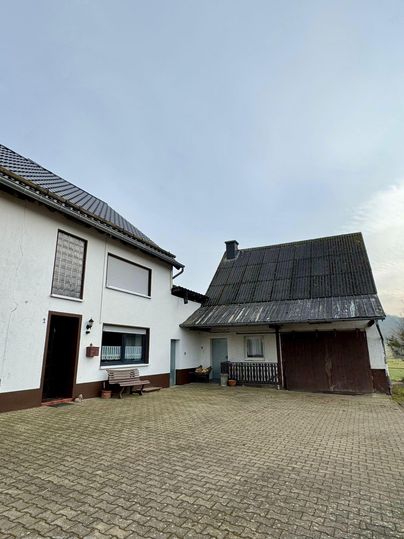 Een eenvoudig huis met een aangrenzend bijgebouw. De binnenplaats is bedekt met bestrating en de lucht is bewolkt.