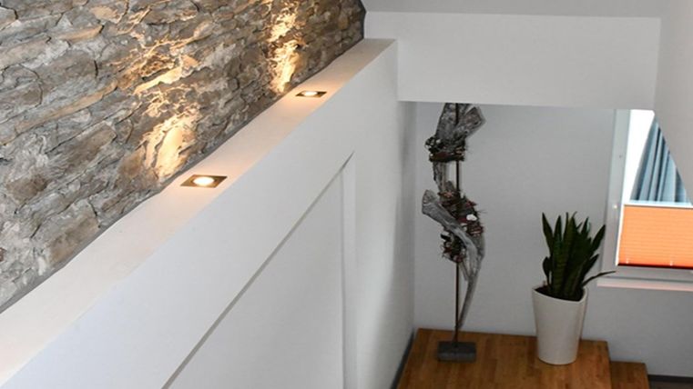 A modern staircase area with a stone wall and stylish lights. Beside the stairs stands a plant in a white pot and a decorative sculpture.