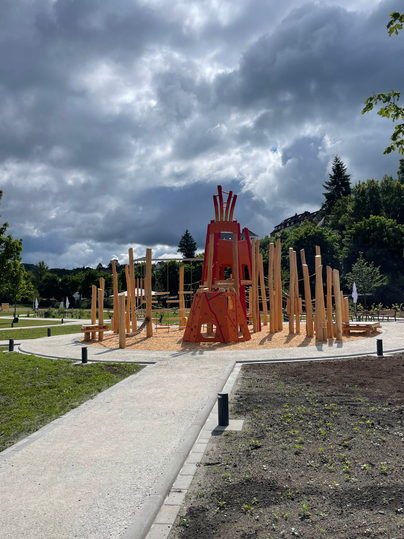 Een nieuwe speeltuin met houten elementen en rode klimstructuren onder een blauwe lucht met wolken. Op de voorgrond is verse opgestorte aarde te zien, daarnaast paden en banken. Verderop zijn er bomen, een kleine schuilplek en een beboste heuvel.
