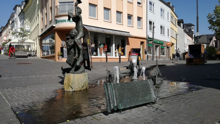 Ein Brunnen mit verschiedenen Skulpturen auf einer Kreuzung. Im Hintergrund Häuser mit Geschäften