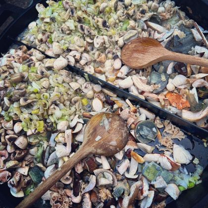 Eine große Pfanne mit einer Mischung aus frischen Meeresfrüchten, Gemüse und Kräutern. Zwei Holzlöffel liegen zum Rühren bereit.