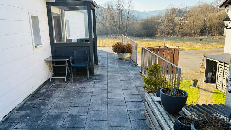 Een terras met een betegelde vloer en een uitzicht op de natuur op de achtergrond. Hier staan enkele planten in potten en een stoel.