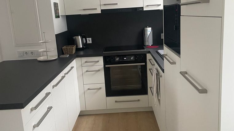 Eine moderne Küche mit weißen Schränken und schwarzen Arbeitsflächen. Der Boden ist aus hellem Holz, und es gibt eine Küchenzeile mit einem Ofen und weiteren Geräten.