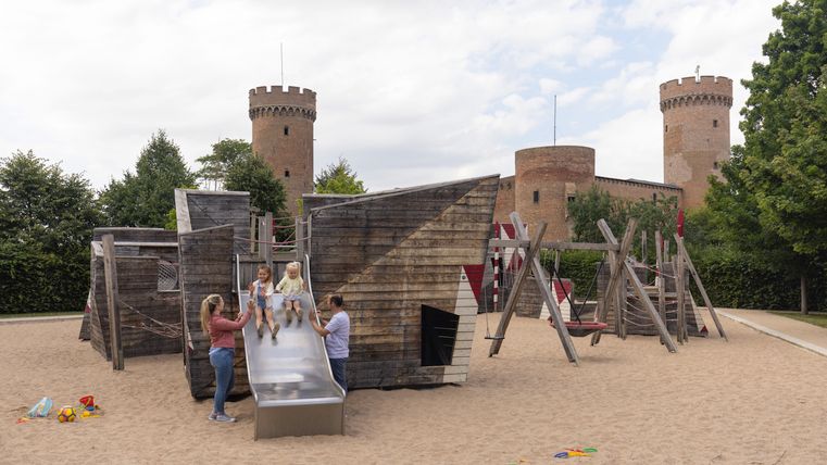 Children play on a playground with a slide and climbing frame in front of a castle backdrop in Zülpich.
