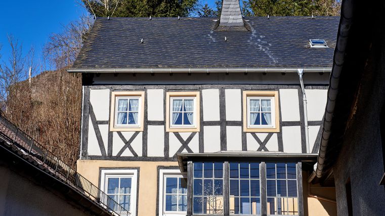 Ein traditionelles Fachwerkhaus mit weißen Wänden und dunklen Holzrahmen. Im Vordergrund sieht man Fenster mit Blick auf die umliegenden Bäume.