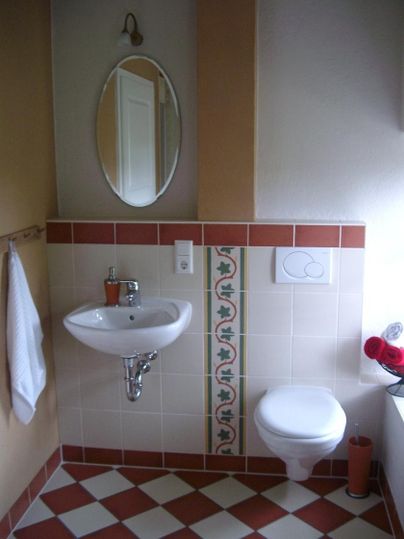 Ein kleines Badezimmer mit einem Waschbecken, einer Toilette und einem runden Spiegel. Der Boden ist mit rot-weißen Fliesen gestaltet und es gibt farbige Handtücher.