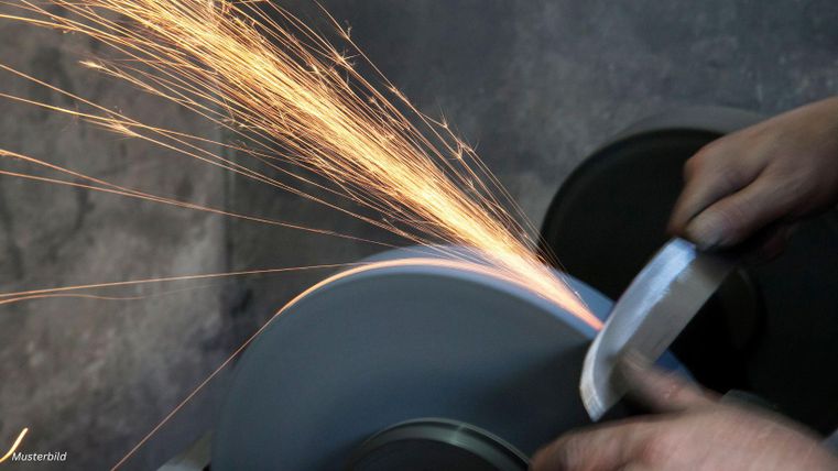 A knife is being sharpened on a grinding stone while sparks fly. Hands are holding the knife firmly.