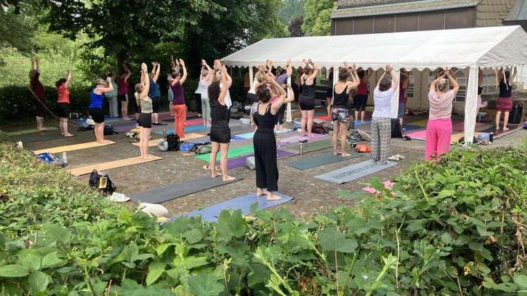 Eine Gruppe von Menschen praktiziert Yoga im Freien unter einem Zelt. Die Teilnehmer haben die Hände über ihren Köpfen und befinden sich auf Matten in einer grünen Umgebung.