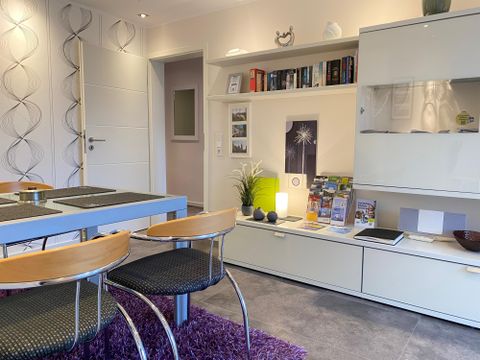 A modern living room with a dining table and chairs. On the wall, there is a shelf with books and decorative elements.