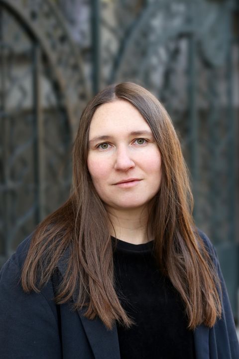 Eine Frau mit langen braunen Haaren steht vor einem historischen Hintergrund. Sie trägt einen schwarzen Pullover und sieht direkt in die Kamera.