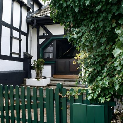 A pretty half-timbered house with green window frames and a sheltered entrance door. The flat garden is surrounded by a green wooden picket fence.