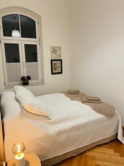 Ein gemütliches Schlafzimmer mit einem Doppelbett und dekorativen Kissen. Der Raum hat große Fenster und eine warme Atmosphäre durch eine Tischlampe.