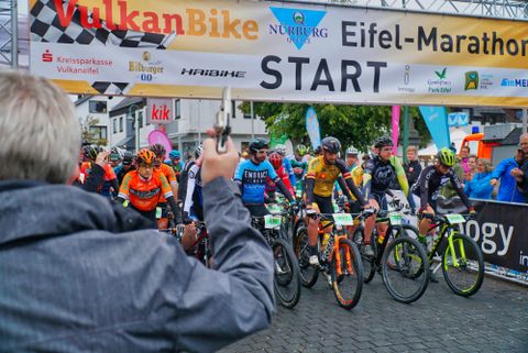 Un groupe de cyclistes se tient au départ du marathon de l'Eifel. En arrière-plan, on peut voir une banderole de départ et des spectateurs attendant la course.