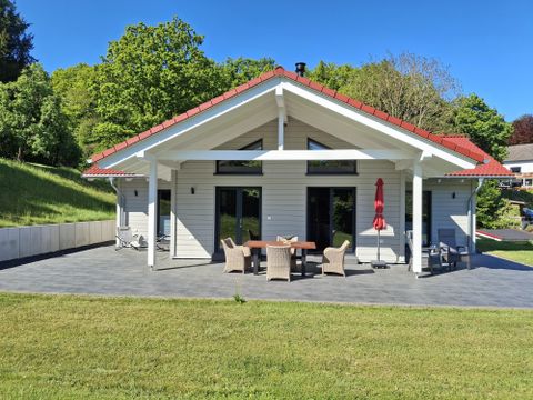 A modern house with a red roof and porch. The garden is green and well-maintained, surrounded by trees.