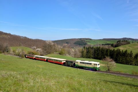A colorful locomotive stands in the snow while people stroll on the platform. The landscape is wintry and picturesque with snow-covered trees.