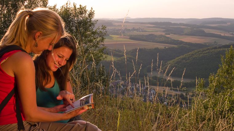 Twee vrouwen zitten op een heuvel en kijken in een boek, op de achtergrond een weids landschap met velden en heuvels.