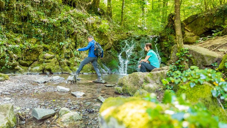 Twee mensen in een bos, de een steekt een beek over, de ander zit op een rots.