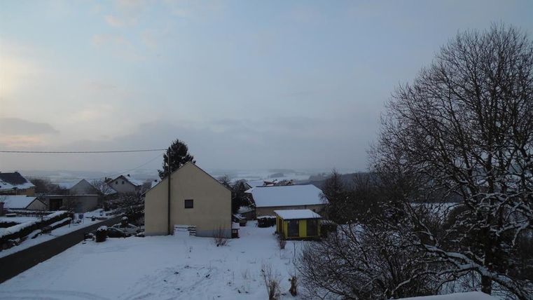 Eine winterliche Landschaft mit schneebedeckten Häusern und Bäumen. Der Himmel ist grau und die Umgebung wirkt ruhig und friedlich.