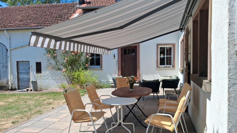 Un espace extérieur confortable avec une table et des chaises sous un store. En arrière-plan, une maison traditionnelle aux murs blancs et aux plantes vertes est visible.